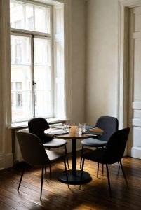 "Calm, neutral mediation meeting room with a round wooden table and three modern dark chairs arranged for discussion. Natural daylight streams through a large window, illuminating water glasses, closed notebooks, and a white tablecloth, creating a professional and welcoming space for family mediation sessions."